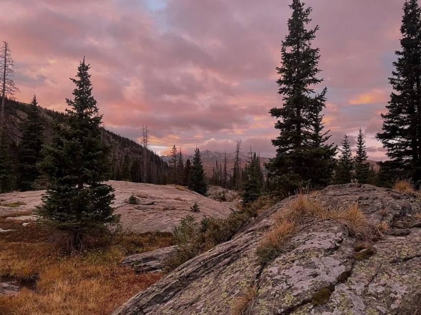 A scenic view of Rocky Mountain National Park at sunrise, featuring rocky terrain and evergreen trees silhouetted against a colorful sky.