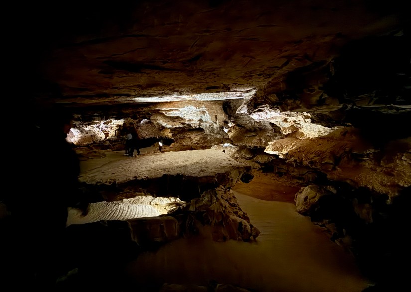 Interior view of a cave with rock formations and soft lighting, showcasing a sandy floor and visitors exploring the space.