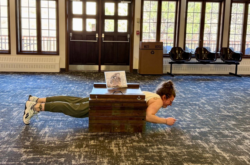 A person lying flat on the floor in front of a wooden box, mimicking a caving position, in a well-lit room with large windows and chairs in the background.