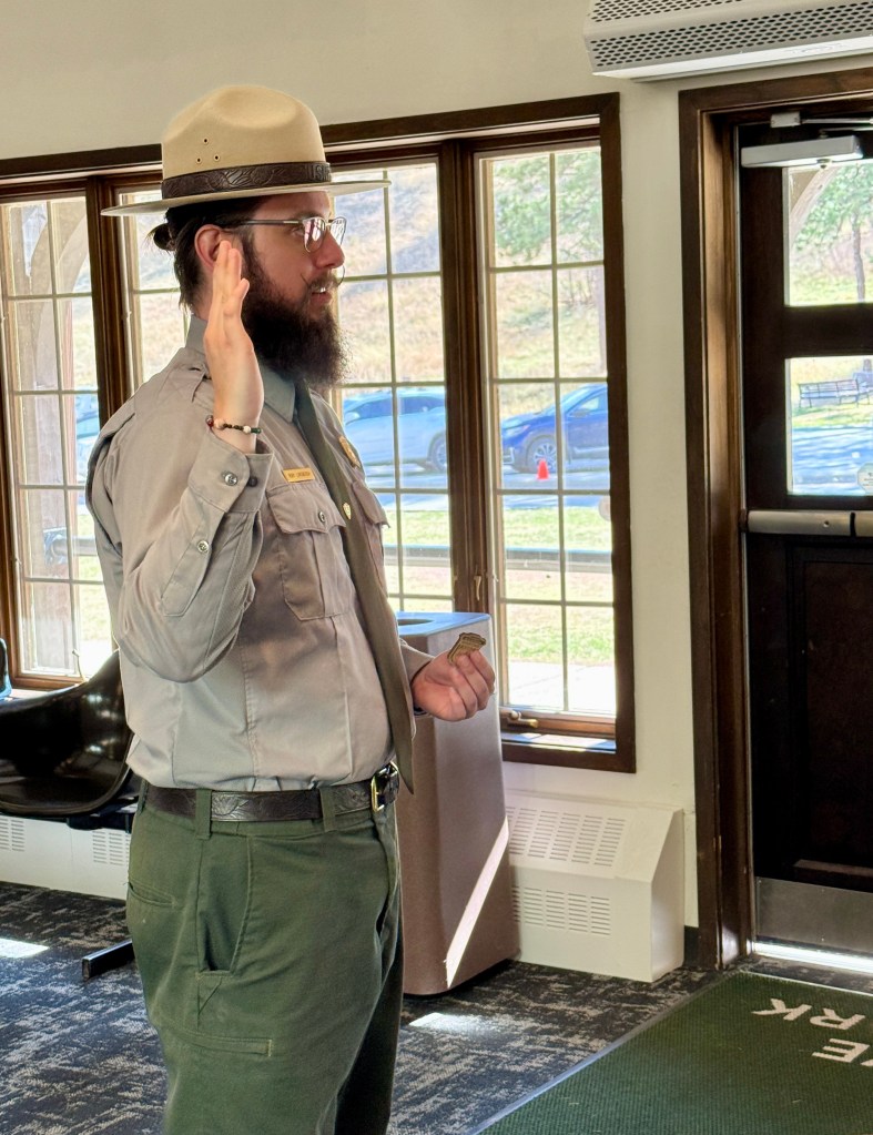 A park ranger in uniform, with a hat and glasses, raises his hand while speaking, standing in a room with large windows and a door in the background.