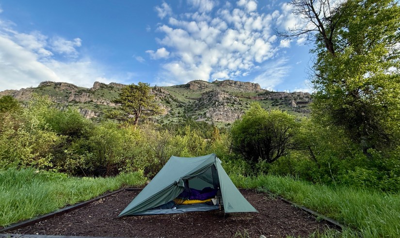 A green tent set up in a natural campsite with lush vegetation and hills in the background under a partly cloudy sky.