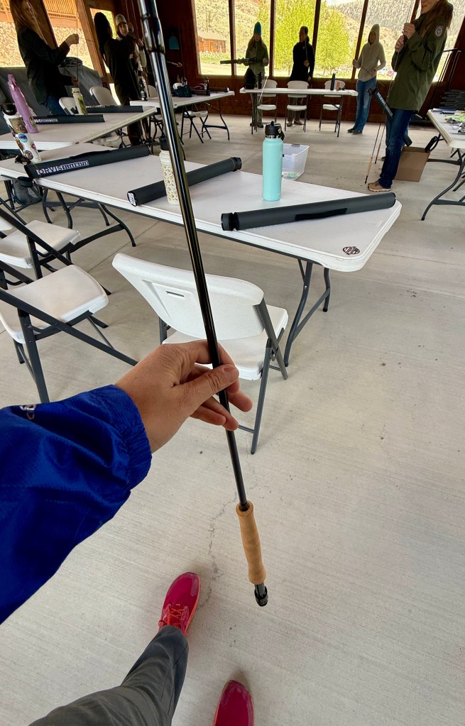 A hand holding a fly rod in a workshop setting with women in the background, showcasing a learning environment for fishing skills.