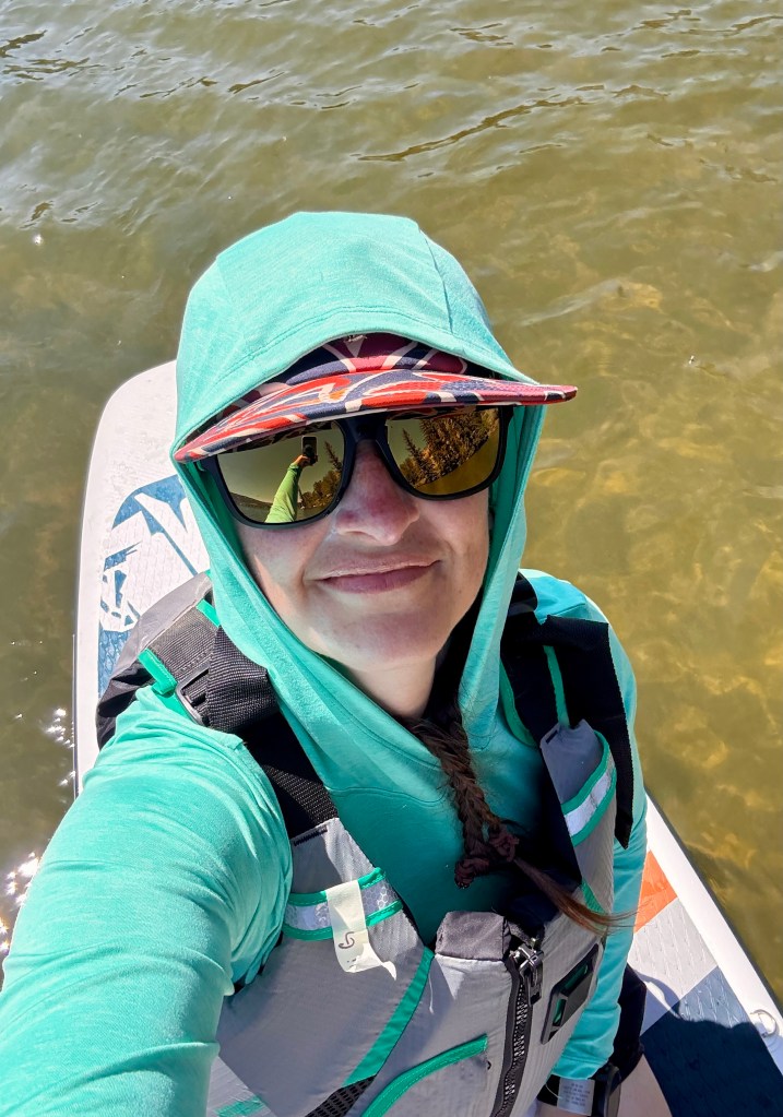 A person wearing a green hooded sweatshirt and sunglasses, smiling while balanced on a stand-up paddleboard in calm water.