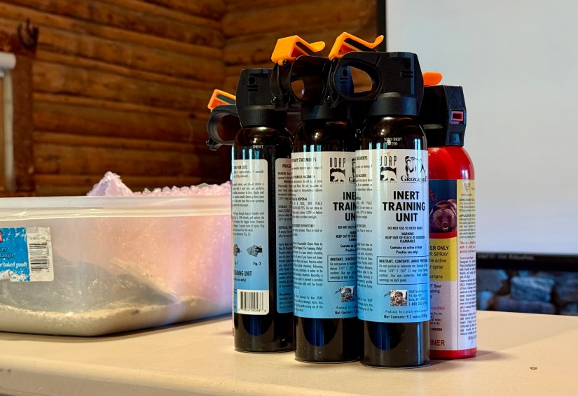 A close-up of several inert bear spray canisters on a table, with a plastic container and a blurred background showing a wooden wall.