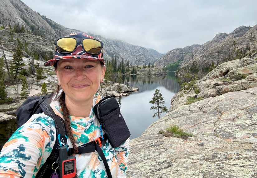 A hiker smiling for a selfie by a tranquil lake surrounded by rugged mountains and trees under a slightly cloudy sky.