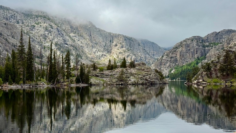 A serene lake surrounded by rugged mountains and lush forests, with reflections of the landscape on the water's surface under a cloudy sky.