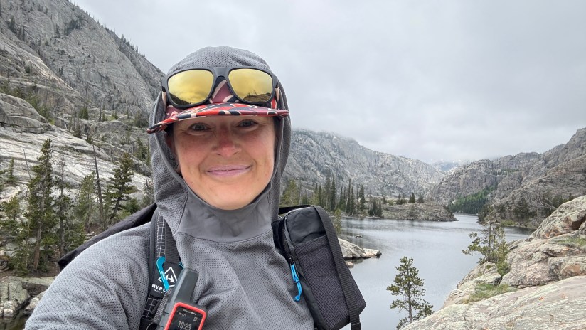 A person wearing sunglasses and a gray hoodie smiles for a selfie in front of a mountainous landscape with a lake, under cloudy skies.