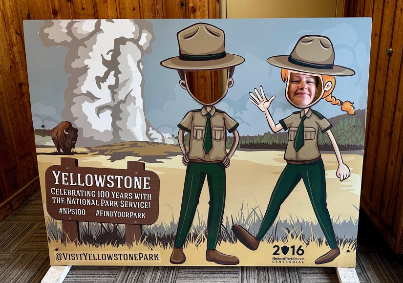 A photo of a playful cutout board featuring ranger characters at Yellowstone National Park, where visitors can pose with their faces in the cutouts. The background depicts a bison and a geyser erupting, celebrating the park's 100-year anniversary.