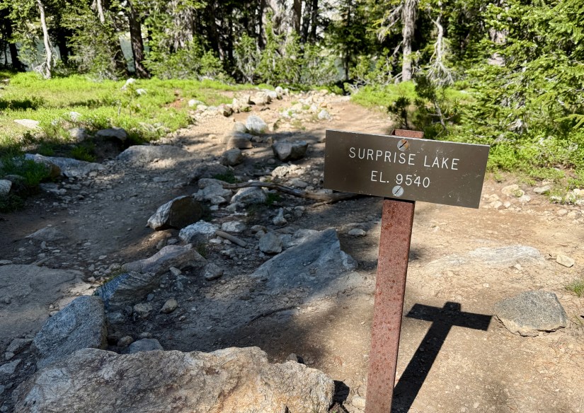 Trail sign indicating Surprise Lake at an elevation of 9,540 feet, surrounded by forested terrain and rocky pathway.