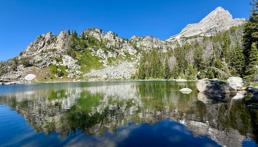 A serene alpine lake reflecting surrounding rocky mountains and forest under a clear blue sky.