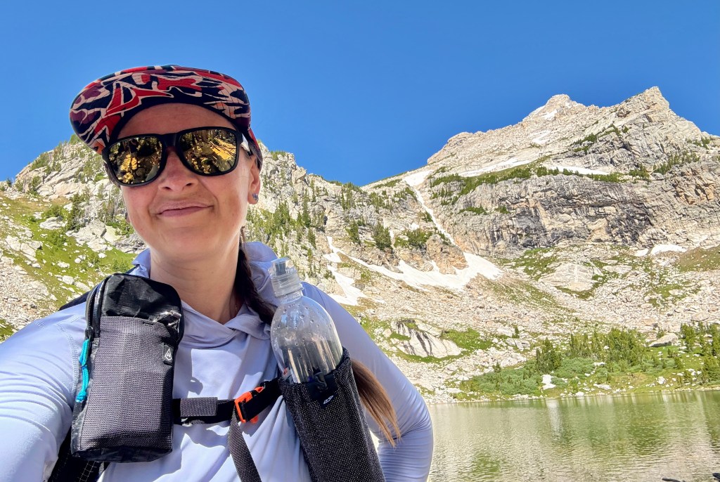 A woman stands in front of a scenic mountain backdrop, wearing sunglasses and a cap, holding a water bottle in a landscape typical of alpine hiking.