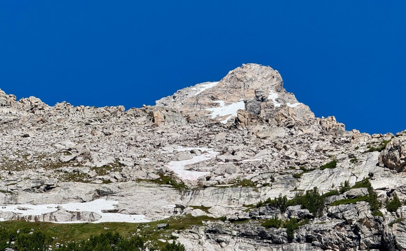 A rocky mountain peak against a clear blue sky, showcasing rugged terrain and patches of snow on the slopes.