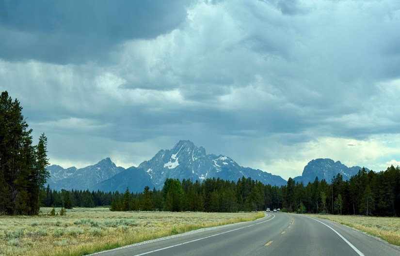 A scenic view of snow-capped mountains looming over a winding road, framed by trees and a cloudy sky.