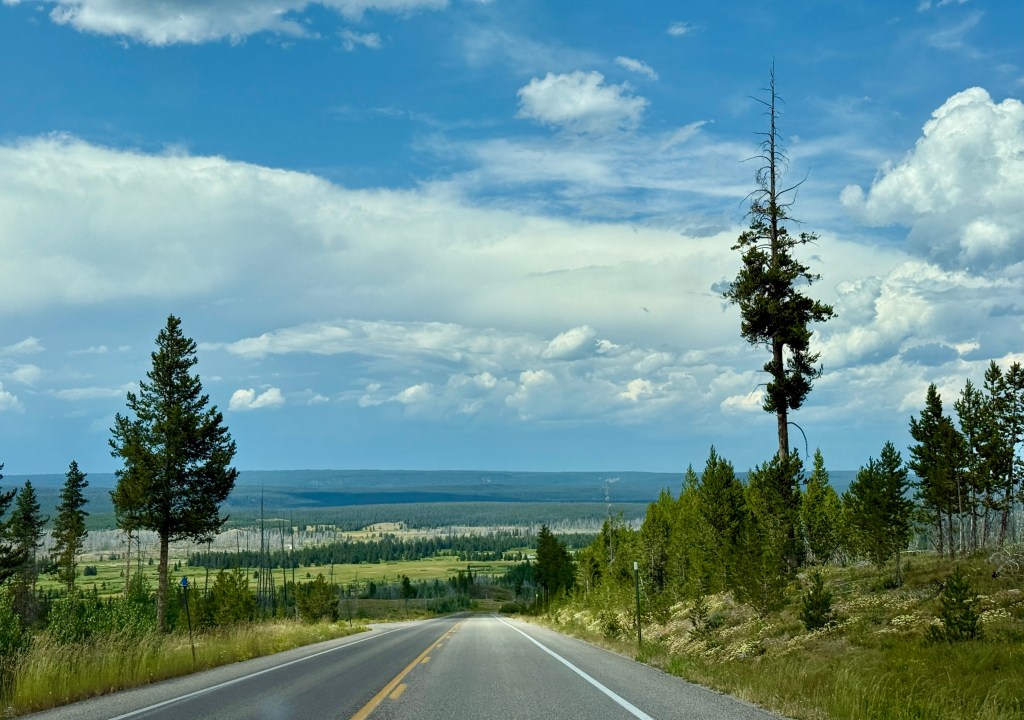 A picturesque view of a winding road through lush green fields and forests, with a bright blue sky dotted with clouds in the background.