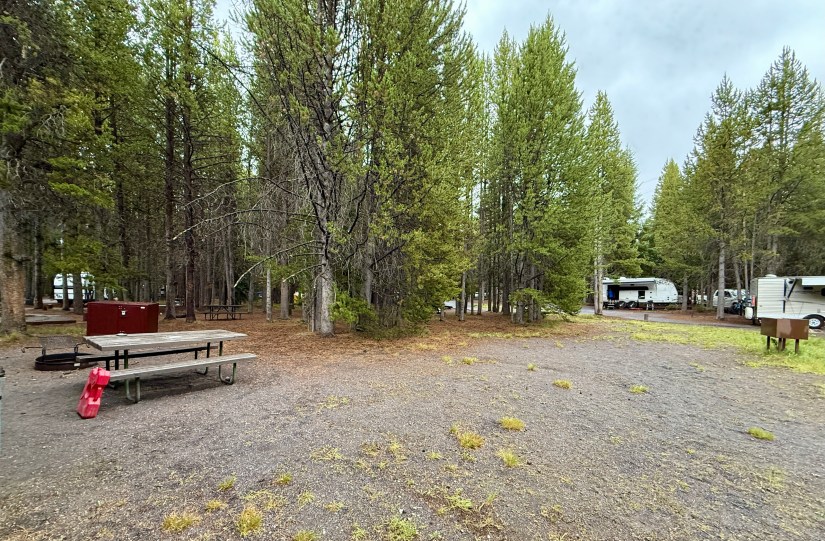 A view of a camp area at Grant Village, Yellowstone National Park, with picnic table, fire pit, and neighboring camp trailers surrounded by tall pine trees and a slightly cloudy sky.