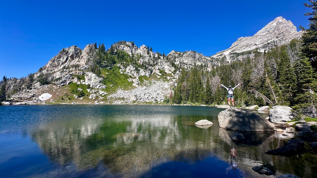 A person stands on a rock by a tranquil alpine lake, surrounded by lush greenery and rugged mountains under a clear blue sky.
