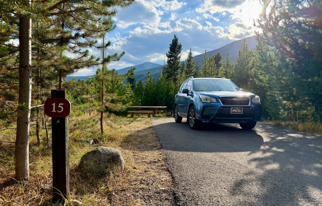 A blue SUV parked near campsite number 15 in a forested area, surrounded by pine trees and mountain views in the background.