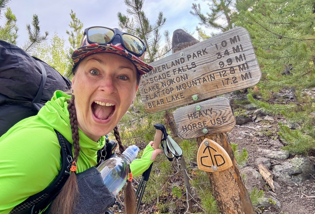 Person with a backpack, smiling and holding a water bottle, stands next to a trail sign indicating distances to various locations in Rocky Mountain National Park.