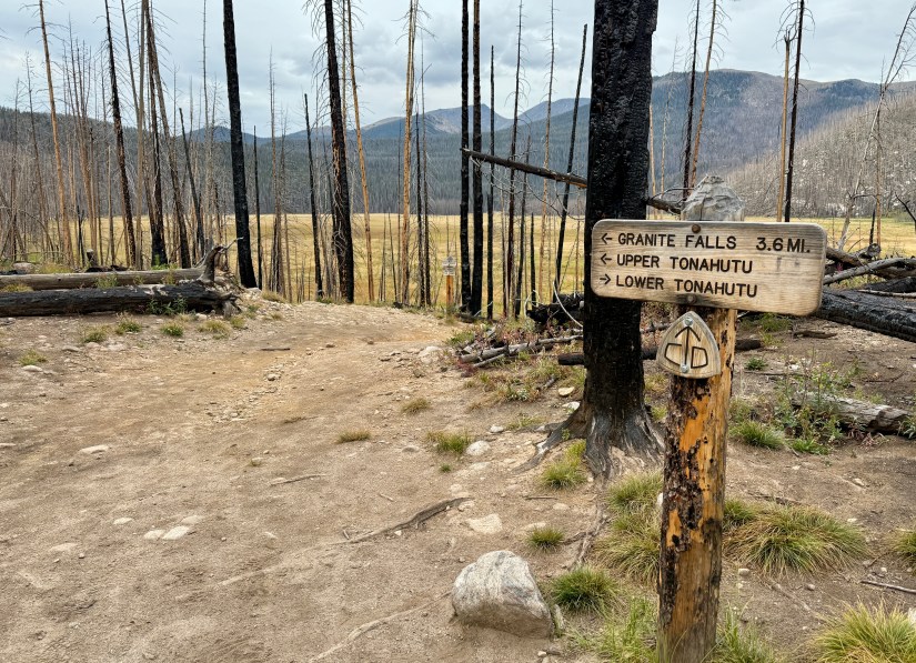 A trail signpost showing directions to Granite Falls, Upper Tonahutu, and Lower Tonahutu, with burnt trees in the background after a wildfire.