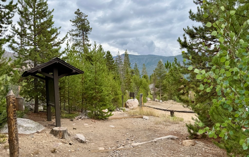 A trailhead shelter surrounded by tall pine trees in Rocky Mountain National Park under a cloudy sky.