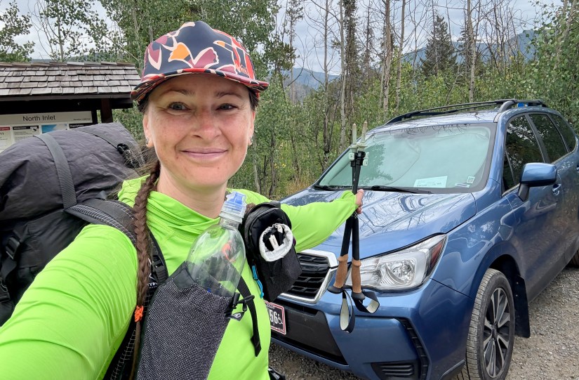 A hiker poses with a backpack and water bottle at North Inlet Trailhead, with a blue SUV parked nearby and trees in the background.