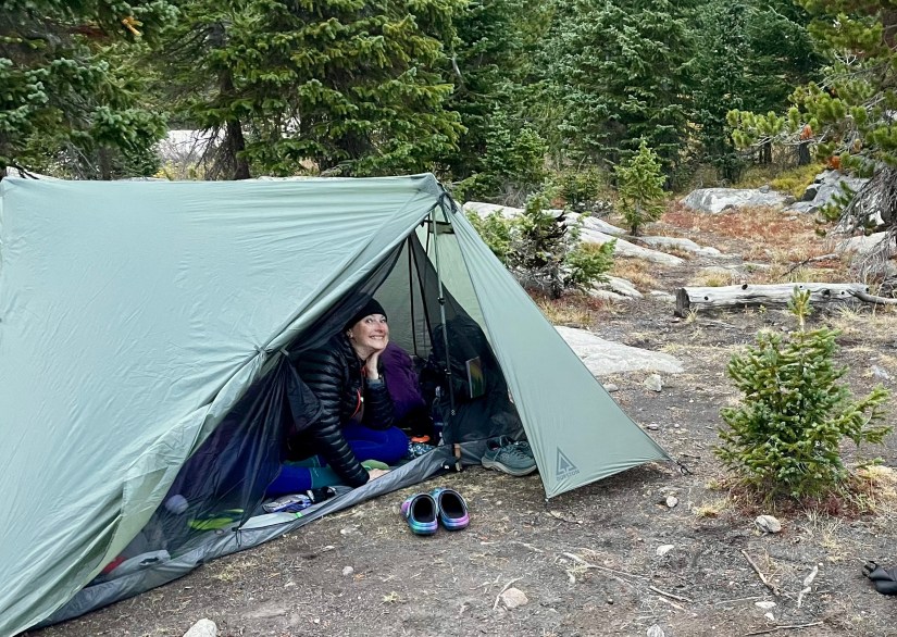 A person sitting inside a tent in a forested area, smiling and looking outside, with hiking gear and shoes visible near the tent entrance.