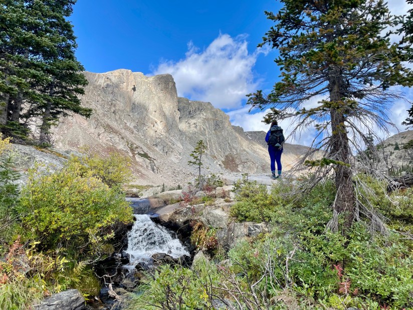 A hiker standing near a small waterfall in a mountainous landscape, surrounded by trees and rocky cliffs under a partly cloudy sky.