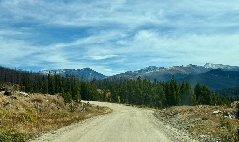 A dirt road winding through a mountain landscape with coniferous trees and rugged peaks in the background, under a blue sky with scattered clouds.