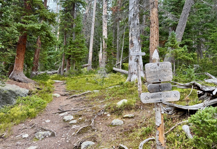 A trail sign indicating directions to Mirror Lake and a no horse policy, surrounded by dense forest in the Rocky Mountain National Park.