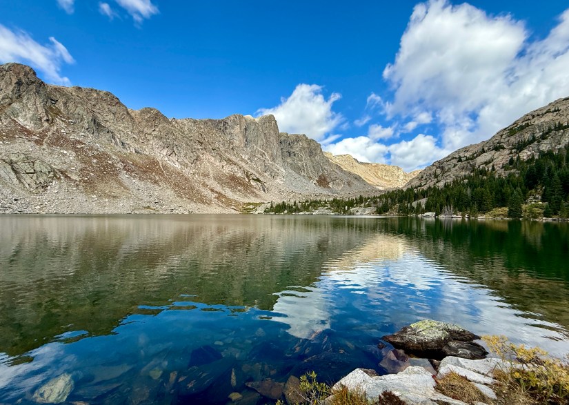 A scenic view of Mirror Lake in Rocky Mountain National Park, surrounded by rugged mountains and lush greenery, with clear blue skies and fluffy white clouds reflecting on the water's surface.