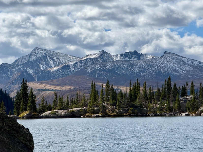 Scenic view of Mirror Lake with snow-capped mountains in the background and pine trees lining the shore under a partly cloudy sky.