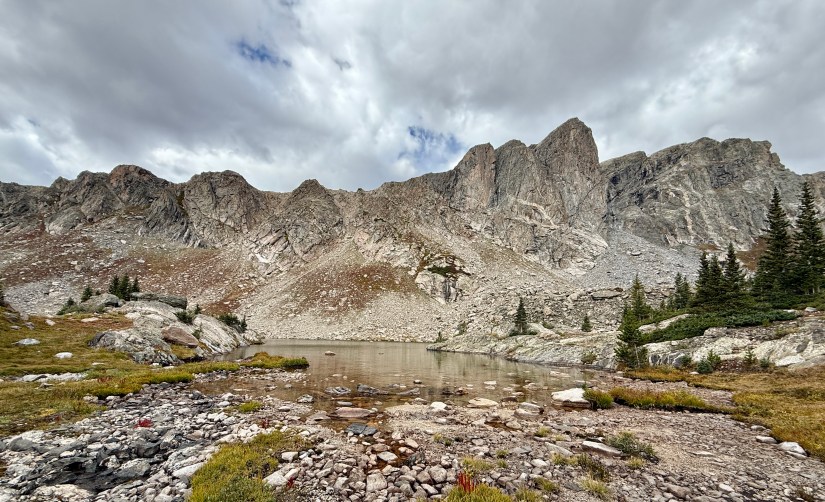 A scenic view of Mirror Lake in Rocky Mountain National Park, framed by rugged granite cliffs and patches of evergreen trees, under a cloudy sky.