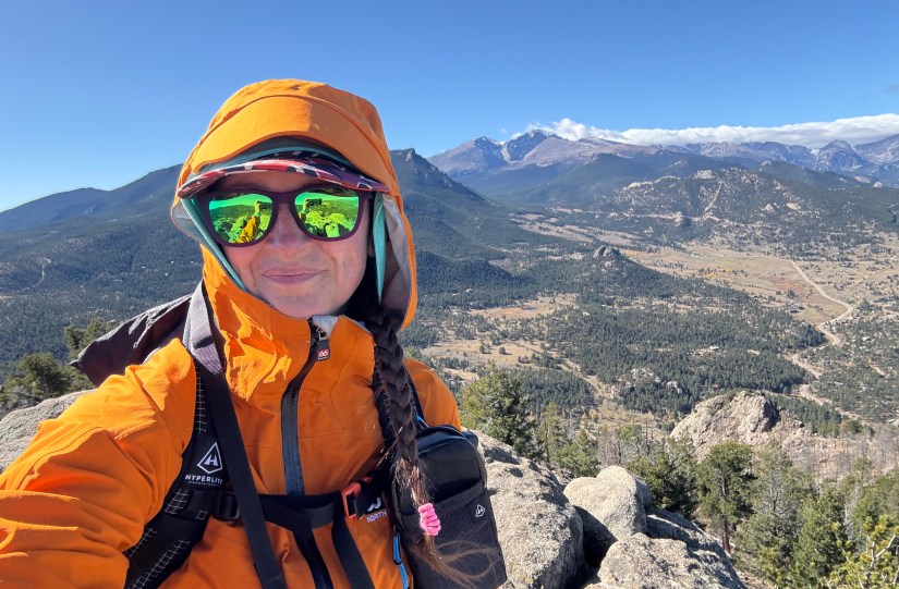 A person wearing an orange jacket and sunglasses poses for a selfie on top of a rocky outcrop, with a panoramic mountain view in the background.