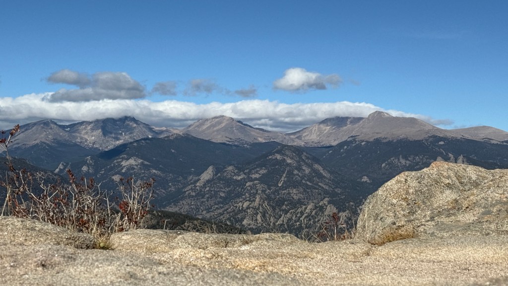 Panoramic view of the Rocky Mountains under a clear blue sky, showcasing rugged peaks and valleys in the foreground.