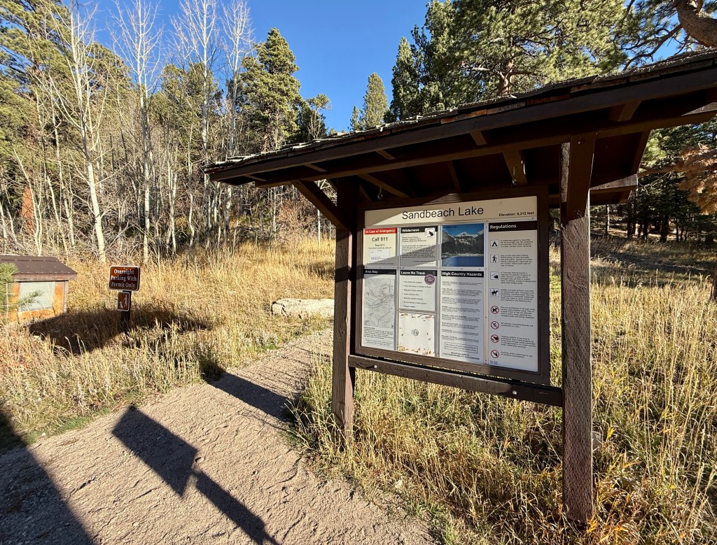 Information sign at the Sandbeach Lake trailhead in Rocky Mountain National Park, surrounded by tall grass and trees.