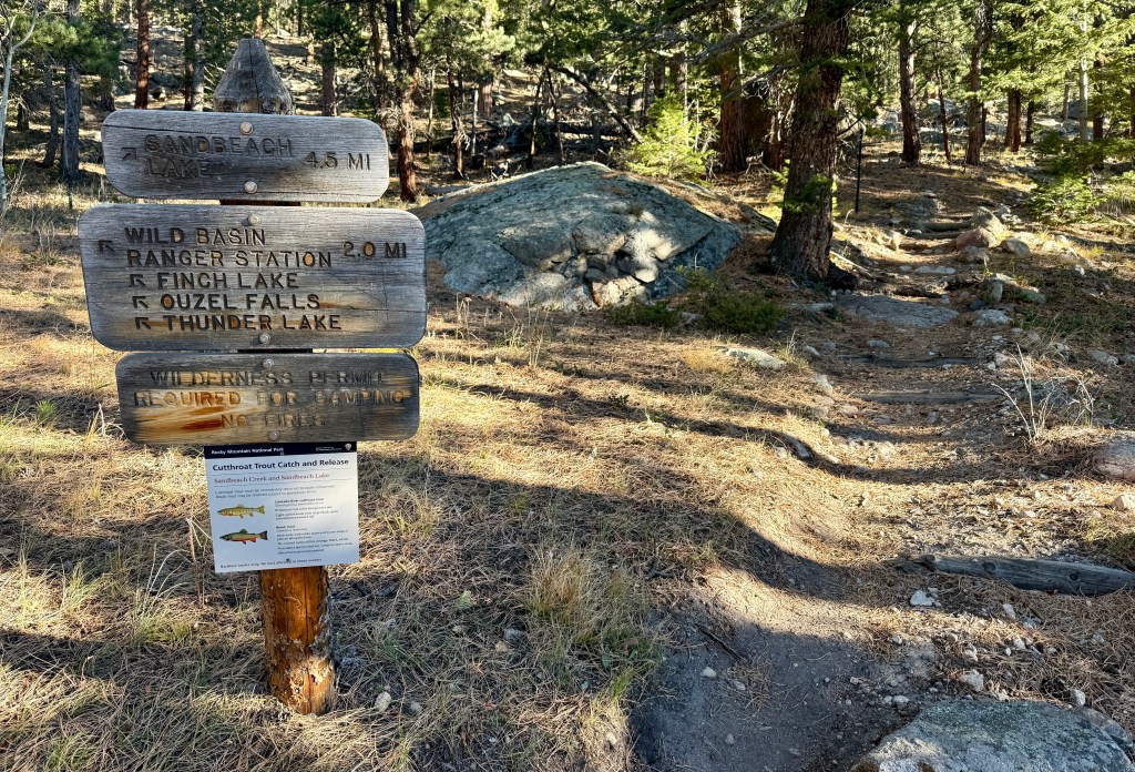 Trail sign indicating directions and distances to Sandbeach Lake, Wild Basin Ranger Station, Finch Lake, Ouzel Falls, and Thunder Lake in Rocky Mountain National Park.