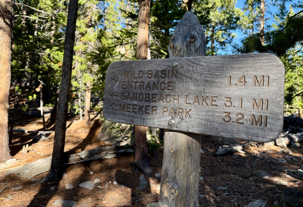 A wooden trail sign indicating directions and distances to Wild Basin, Sandbeach Lake, and Meeker Park surrounded by trees in a forested area.
