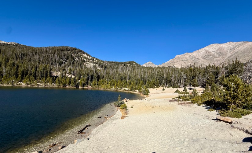 A scenic view of Sandbeach Lake in Rocky Mountain National Park, featuring a sandy shore, clear blue water, and surrounding pine forests under a bright blue sky.