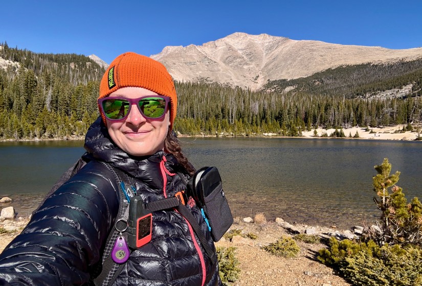 A person wearing a bright orange beanie and sunglasses stands in front of a mountain backdrop beside a tranquil alpine lake, dressed in a black puffy jacket.
