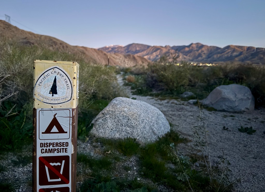 A signpost marking the Pacific Crest Trail, indicating a dispersed campsite and water restrictions, with mountains in the background under a morning sky.