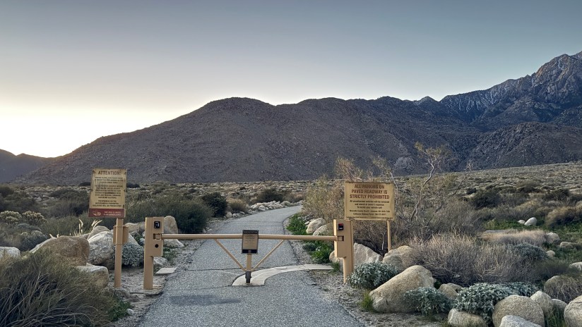 A gated entrance to a desert trailhead with signage indicating restrictions, surrounded by a mountainous landscape during early morning light.