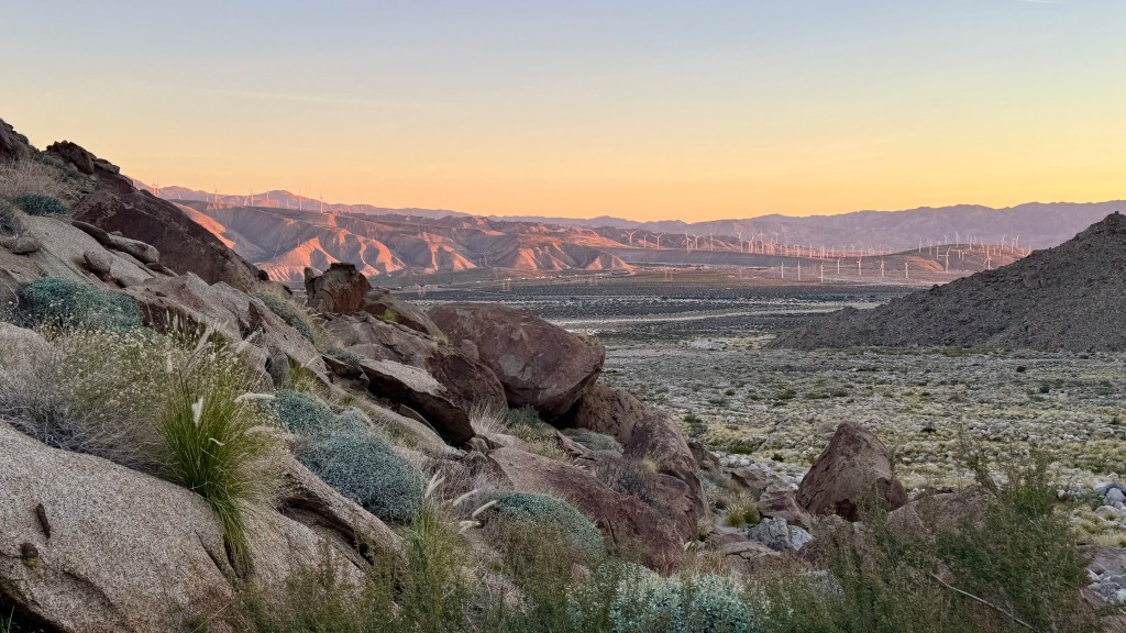 Scenic view of rocky terrain and desert landscape at sunrise, with mountains in the background and wind turbines on the horizon.