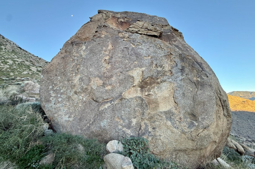 A large boulder situated in a desert landscape, with grassy vegetation at its base and mountains visible in the background. The sky is clear with a hint of moonlight.