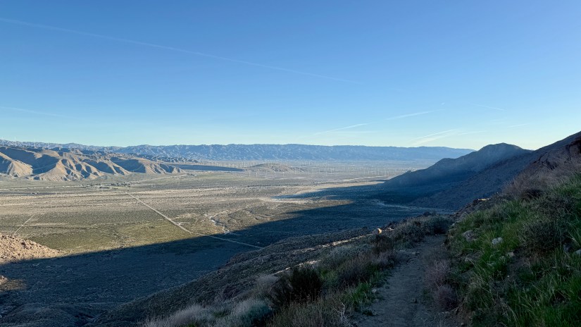A panoramic view of the desert landscape with mountains in the background, showcasing a clear blue sky and a hiking trail along the ridge.