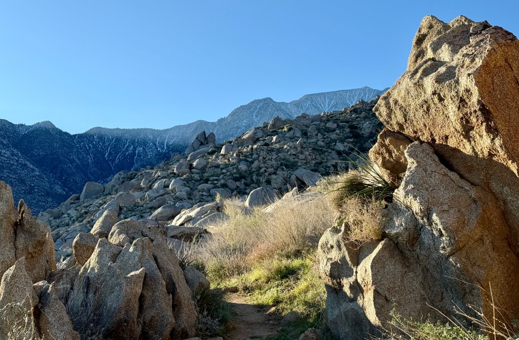 A scenic view of rugged mountain terrain with large boulders in the foreground, featuring a clear blue sky and snow-capped mountains in the background.