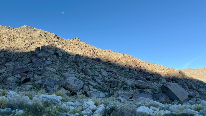 Rocky terrain of a desert mountain landscape under a clear blue sky with a visible crescent moon.