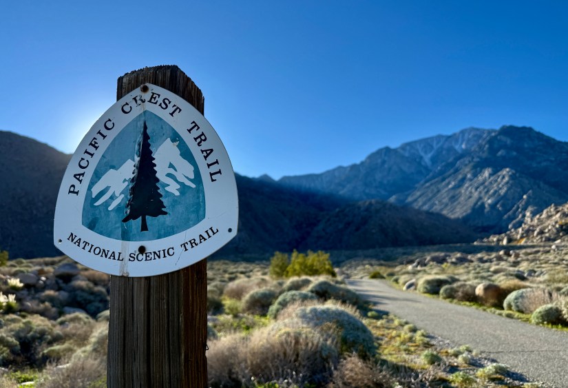 Sign marking the Pacific Crest Trail with a mountainous background and clear blue sky.