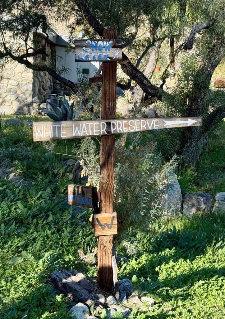 Wooden signpost indicating directions to Snow Creek and White Water Preserve, surrounded by greenery.
