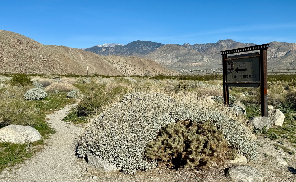 A sign for the Pacific Crest Trail (PCT) points the way towards Big Bear, surrounded by desert vegetation and mountains in the background.