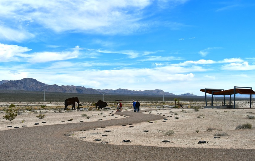 Wide view of a desert landscape with two mammoths and visitors walking along a path, against a backdrop of mountains and a blue sky.
